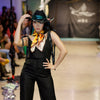 Model on a runway wearing a black outfit with a colorful scarf, surrounded by an audience.