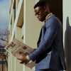 Man in a blue suit reading a newspaper against a building.